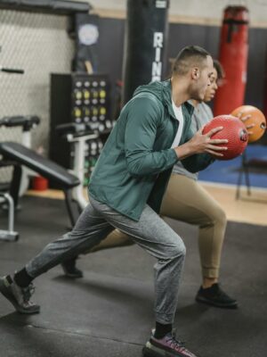 Side view of sporty young multiethnic male trainer and female client doing forward lunges exercise with medicine ball during workout in gym together