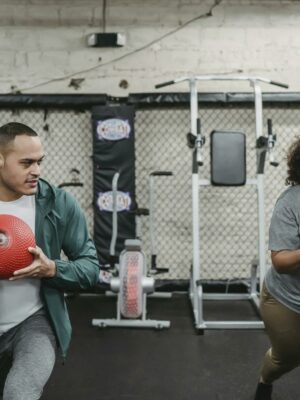 Young muscular ethnic male instructor in activewear doing lunges exercise with ball near plus size black woman during training in sport club